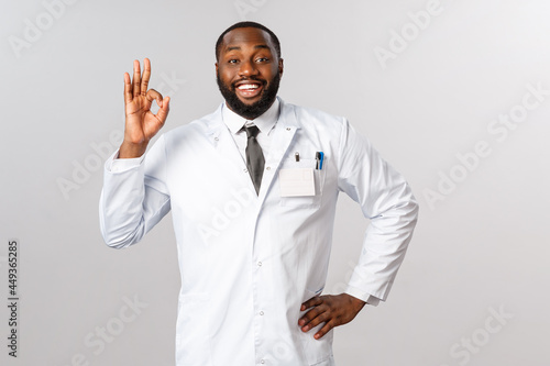 Portrait of handsome optimistic african-american male doctor guarantee you be fine during coronavirus oubreak pandemic if staying home, social-distancing and avoiding crowd places, show ok sign photo