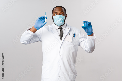 Covid19, pandemic and healthcare concept. Cheerful handsome african-american doctor showing vaccine in ampule and syringe, ready to tream patients, prepared for hospital check-up, grey background photo