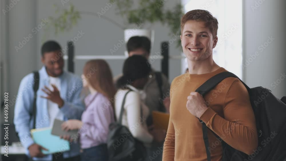 Confident guy with backpack smiling to camera, posing at college over chatting diverse groupmates on background