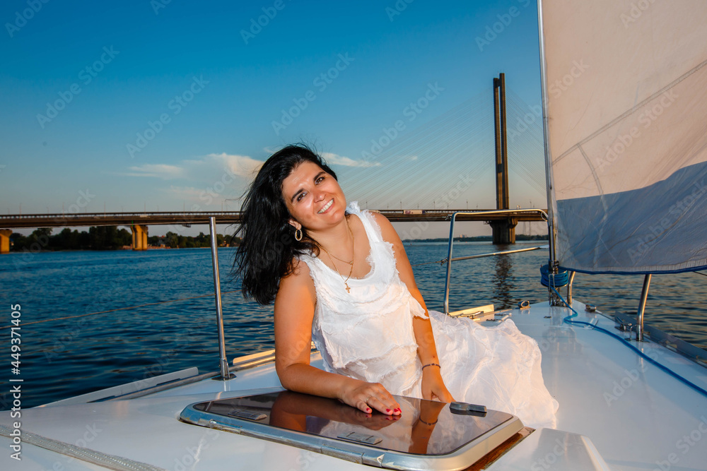 portrait of a happy toothy smiling beautiful girl with a white dress and curly dark hair, standing on a yacht in the summer and in the blue sky.