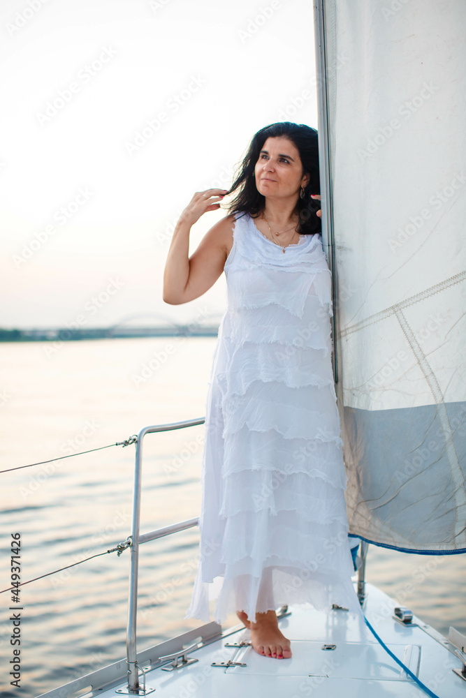 portrait of a happy toothy smiling beautiful girl with a white dress and curly dark hair, standing on a yacht in the summer and in the blue sky.