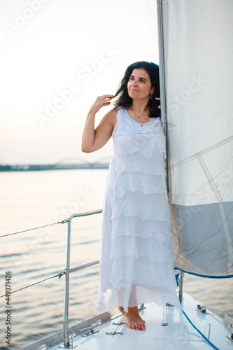 portrait of a happy toothy smiling beautiful girl with a white dress and curly dark hair, standing on a yacht in the summer and in the blue sky. © popovatetiana
