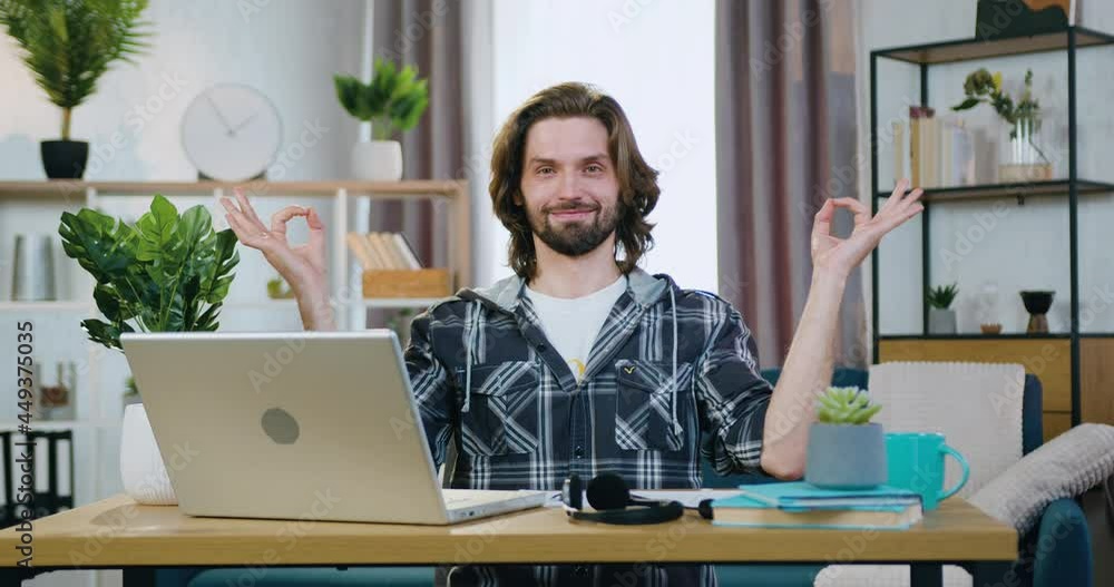 Attractive smiling peaceful young bearded man relaxing and making gyan mudra symbols when meditating at his workplace during working on computer at home Attractive smiling peaceful young bearded man relaxing and making gyan mudra symbols when meditating at his workplace during working on computer at home