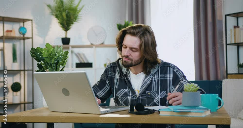 Remote work concept where attractive confident serious skilful young bearded man sitting at the desk at home and working with computer and papers Remote work concept where attractive confident serious skilful young bearded man sitting at the desk at home and working with computer and papers