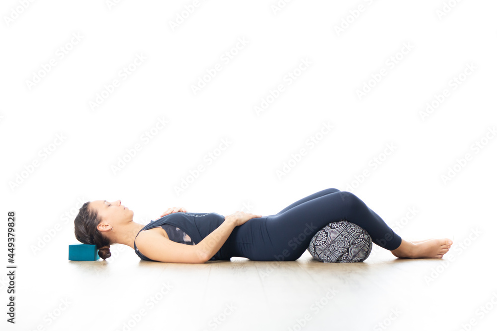 Restorative yoga with a bolster. Young sporty attractive woman in bright white yoga studio, lying on bolster cushion, stretching and relaxing during restorative yoga. Healthy active lifestyle
