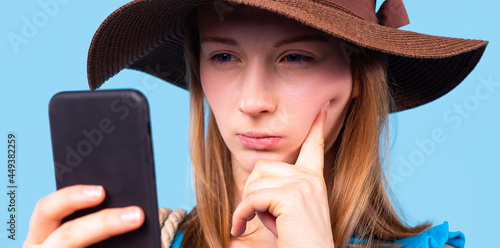 Close up photo of a pensive blonde girl with squints eyes wearing the brown hat looking at the phone holding in her hand.  photo