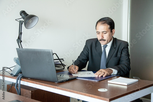 Middle age old business executive with moustache wearing suit and tie writing with pen at his office workstation.
