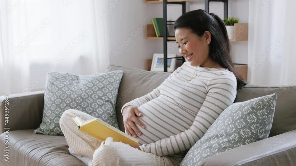 pregnancy, people and leisure concept - happy smiling pregnant asian woman sitting on sofa and reading book at home pregnancy, people and leisure concept - happy smiling pregnant asian woman sitting on sofa and reading book at home