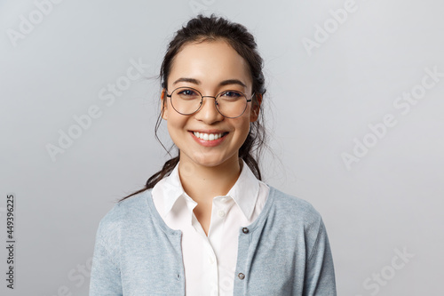 Close-up portrait of attractive, friendly-looking asian female office worker, employee or teacher in glasses, smiling broadly camera with enthusiastic attitude, stand grey background