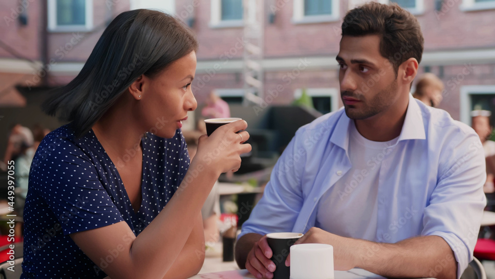 Attractive diverse young couple having lunch sitting at cafe table outdoors drinking coffee talking