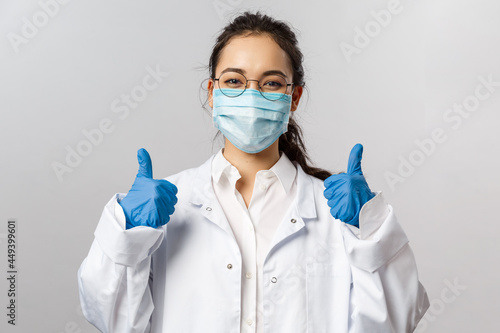 Doctors, infectionist, research and covid19 concept. Satisfied young asian female doctor receive good results on studying virus, discover vaccine, show thumb-up, wear face mask and gloves photo