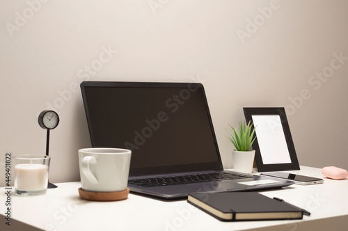 Picture of laptop on a white table with a mug a clock a small bush a notebook a phone a picture frame and a headphone case