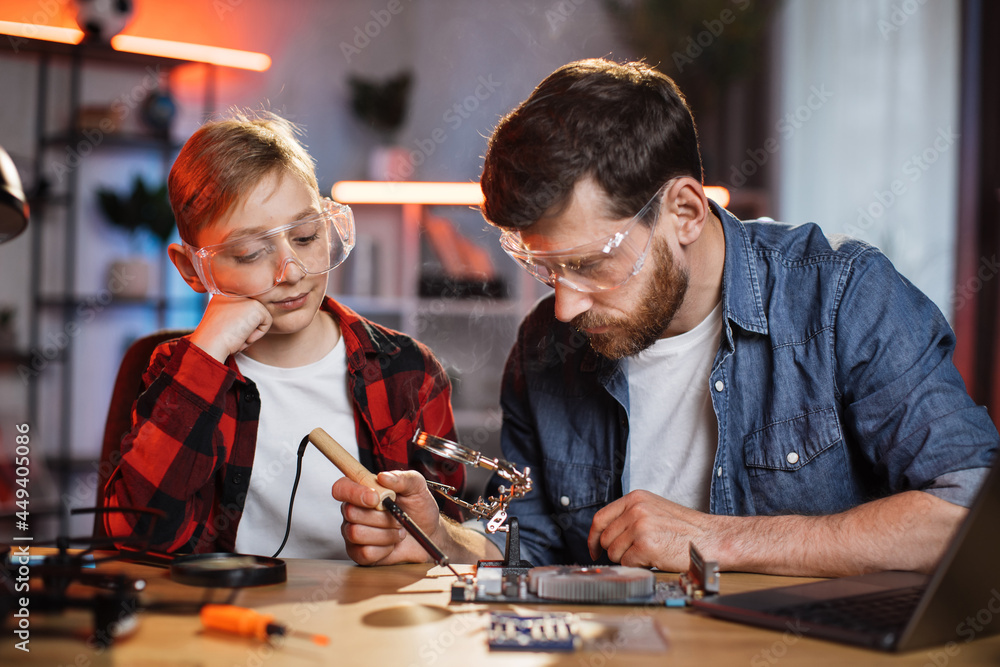 Cute little boy in protective glasses and casual wear sitting at tablet near his father that soldering display card from laptop. Father and son spending time usefully at home.