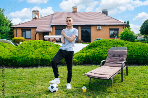 Full length young caucasian man with amputated arm, holds foot on soccer ball while standing on the lawn in the backyard of his home, disability rehabilitation and active lifestyle