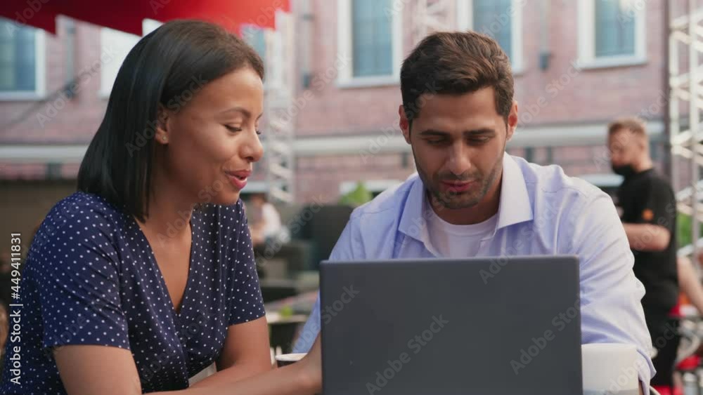 Multiethnic couple of colleagues talking and working on laptop together sitting in cafe outdoors Multiethnic couple of colleagues talking and working on laptop together sitting in cafe outdoors
