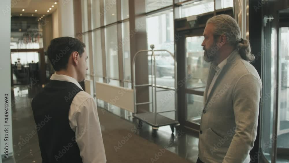 Slowmo tracking shot of mature businessman with suitcase walking into modern hotel being welcomed by young male receptionist in uniform Slowmo tracking shot of mature businessman with suitcase walking into modern hotel being welcomed by young male receptionist in uniform