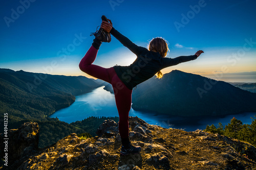 Adventurous athletic woman hiker standing in a yoga pose on a mountain top with a sunset over mountains and a beautiful lake.
