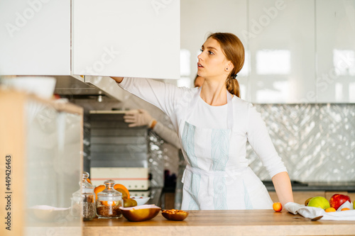 Young attractive adult woman wearing apron cleaning the kitchen after cooking, wipes the table with wiper at cozy home. Adult daughter and her good looking middle aged mother preparing food together.