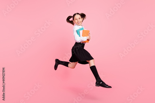 Full length photo of young cheerful small girl jump up hold copybooks smile long socks isolated on pink color background © deagreez