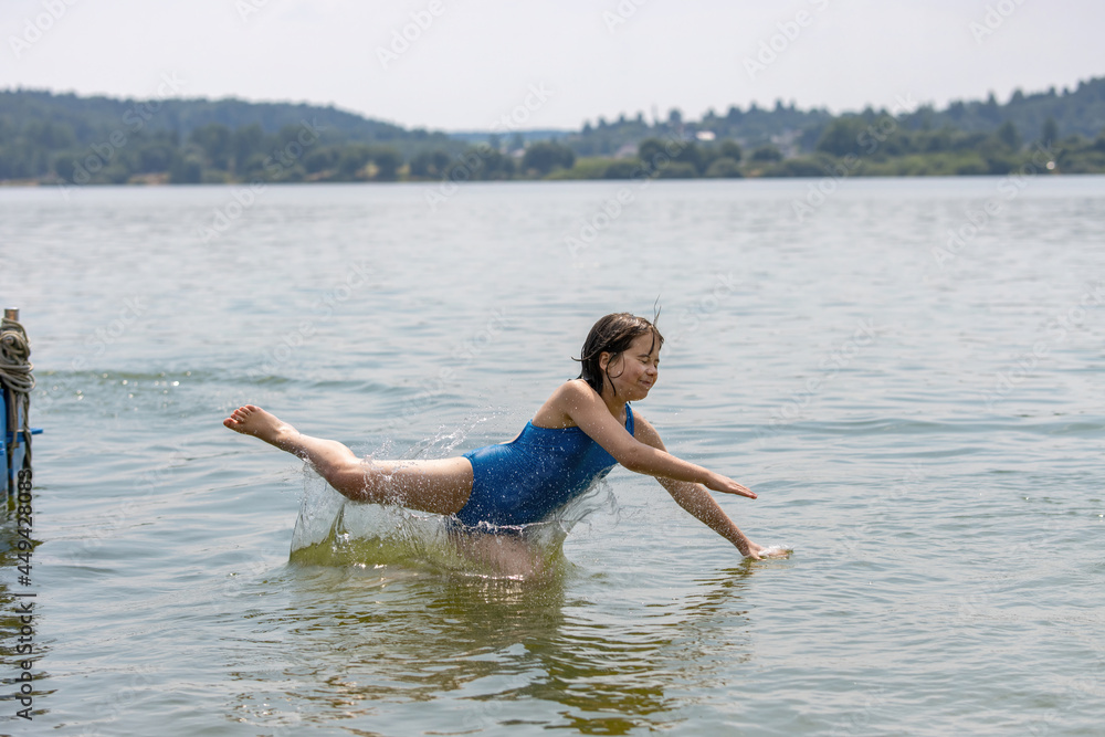 Teenage girl in blue swimsuit bathes in calm cool lake water in hot summer, raising a lot of splashes