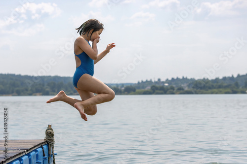 Fototapeta Naklejka Na Ścianę i Meble -  A teenage girl in a blue swimsuit jumps from a pier into the calm cool lake water in a hot summer