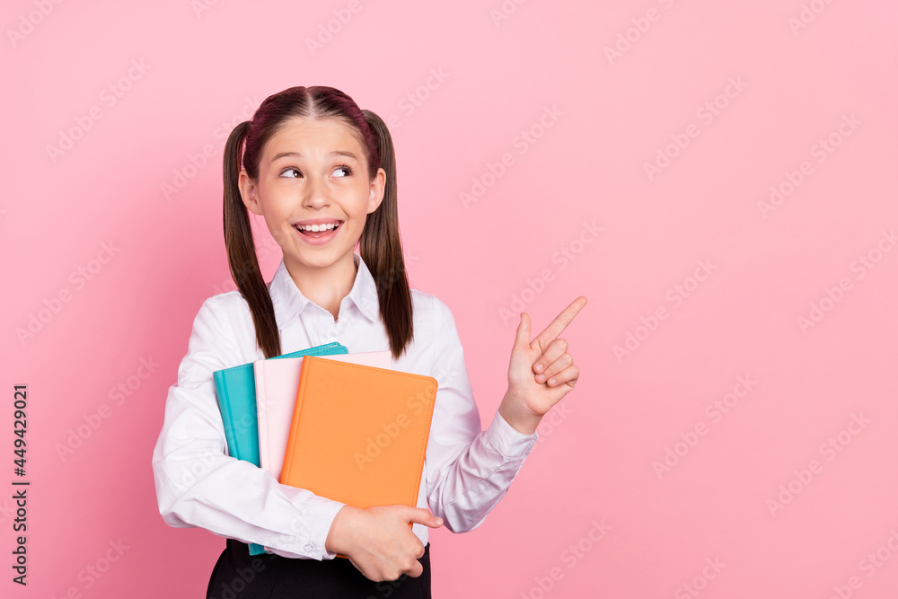 Photo of young happy positive small girl point finger empty space smile hold notes isolated on pink color background Photo of young happy positive small girl point finger empty space smile hold notes isolated on pink color background