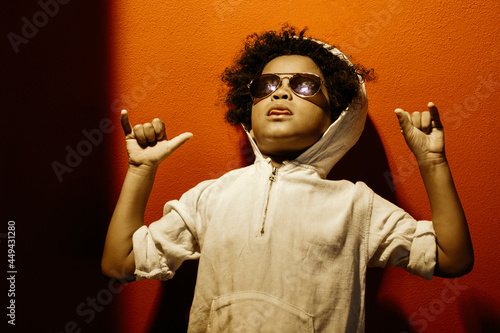 Black boy, with afro hair and sunglasses. With a cocky and funny expression like a rapper. With a red wall in the background. Children, future, education, immigration, and equality concept.