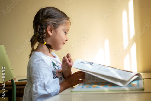 Adorable niña en su primer día de colegio  vestida con mandilón a cuadros verdes dibujando y mirando libros.