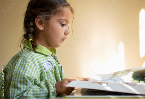 Adorable niña en su primer día de colegio  vestida con mandilón a cuadros verdes dibujando y mirando libros.
