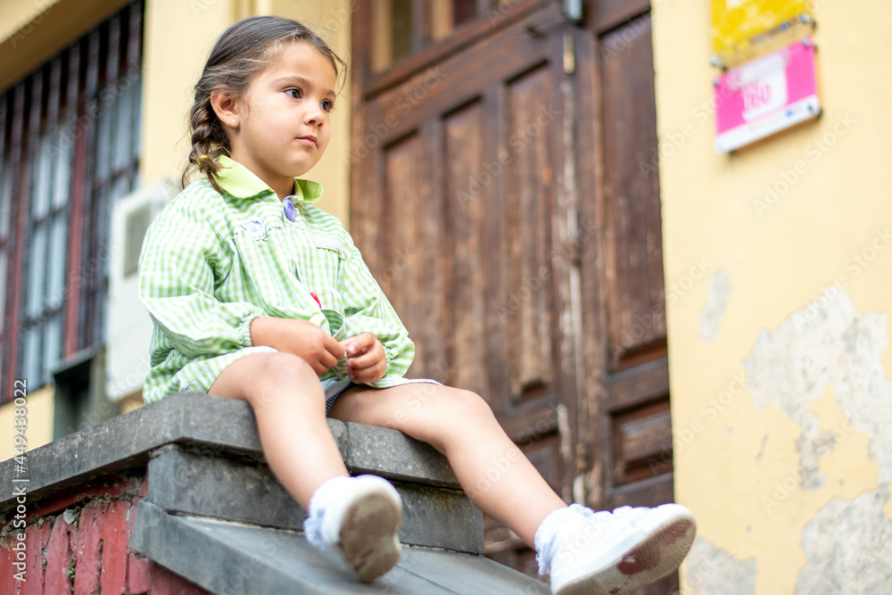 Adorable niña en su primer día de colegio  vestida con mandilón a cuadros verdes dibujando y mirando libros.