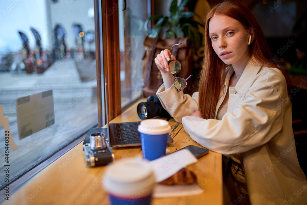 Confident redhead female photographer with modern laptop sitting at cafeteria table and thinking about distance job, contemplative woman with netbook device pondering on idea for web design