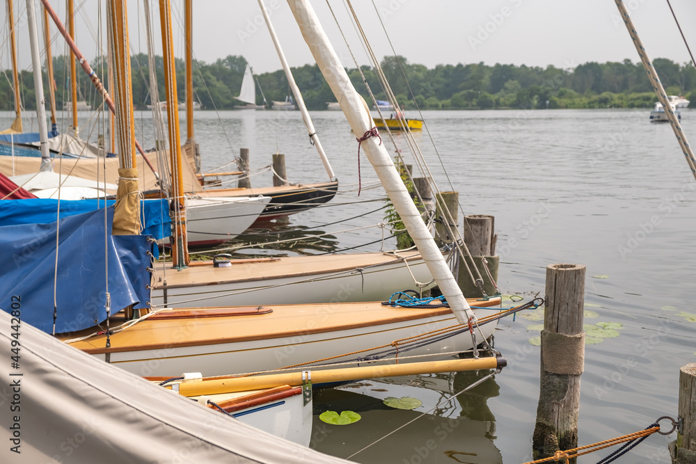 Fototapeta premium Wroxham, Norfolk Broads, UK – July 2021. Sailing boats moored along the wooden staging on the side of Wroxham Broad during the annual sailing regatta open week.