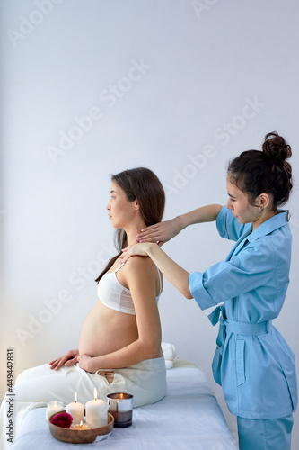 Beautiful gravid asian brunette woman enjoying back and shoulders massage in beautician room in spa center, side view on relaxed lady sitting on bed, having rest. female physiotherapist doing massage