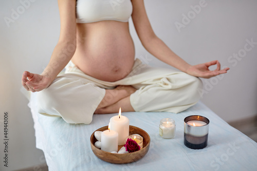 Gravid Young Female Practices Yoga In Bright Room, Health During Pregnancy. Mom-to-be In Casual Outfit Sitting With Crossed Legs, Meditating. Motherhood, Harmonious Pregnancy. Copy Space photo