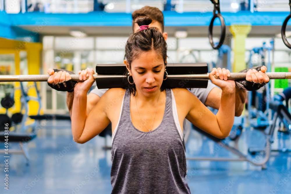 trainer putting a weighted shoulder bar and pad on a young Hispanic Latina woman shoulder to lift weights trainer putting a weighted shoulder bar and pad on a young Hispanic Latina woman shoulder to lift weights