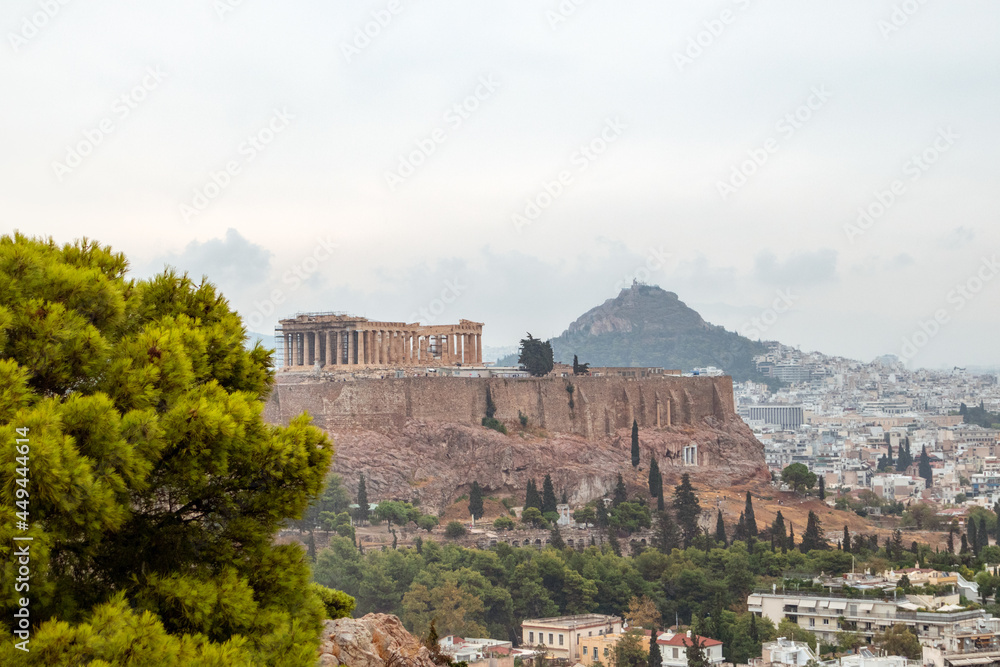 Obraz premium Acropolis (Parthenon, Temples), Mount Lycabettus and white city buildings with vivid pine summer greenery. Athens ancient historical landmark from Filopappou Hill on cloudy day
