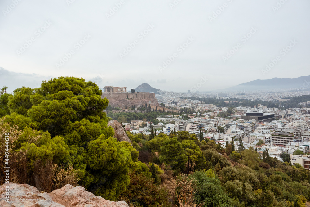 Obraz premium Acropolis (Parthenon, Temples), Mount Lycabettus in distance and white city buildings with vivid pine greenery. Athens historical landmark from Filopappou Hill on cloudy day