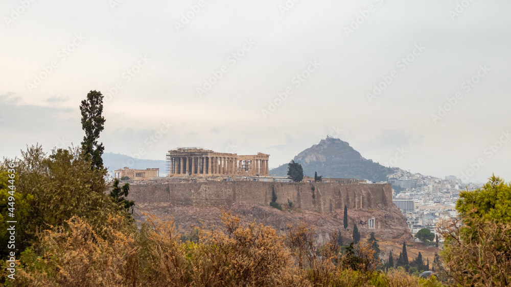 Fototapeta premium Acropolis (Parthenon, Propylaea, Temples, Odeon of Herodes Atticus), Mount Lycabettus and white city buildings in summer bushes. Athens ancient historical landmark from Filopappou Hill on cloudy day