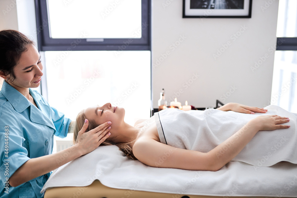 Fototapeta premium woman receiving osteopathic or chiropractic treatment on face and head in clinic, side view on female lying on bed getting massage procedure by woman, relaxing with eyes closed, side view portrait