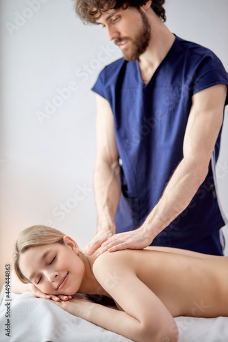 Portrait Of Bearded Nice Masseur In Uniform Carefully Doing Massage On Female Back, Blonde Caucasian Woman Lying On Belly Relaxing, Enjoying. Copy Space For Advertisement. Focus On Woman