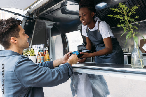 Young man with credit card paying to a saleswoman at a food truck. Female entrepreneur in apron receiving payment from a customer.