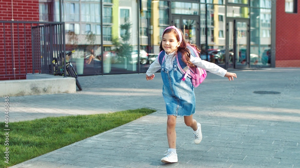 Portrait of Caucasian cute happy little school girl with pink backpack running on schoolyard after lessons. Pretty joyful small female junior student smiling outdoors on street. Emotions concept Portrait of Caucasian cute happy little school girl with pink backpack running on schoolyard after lessons. Pretty joyful small female junior student smiling outdoors on street. Emotions concept