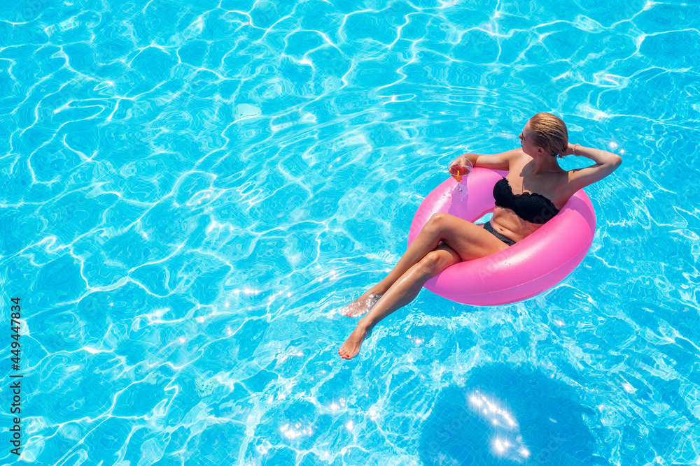 A young woman swims on a pink inflatable circle in clear blue water. A girl with a cocktail relaxes in the pool on a sunny day