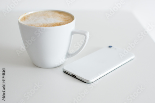 A white smartphone lies on a white table against a white wall. In the background a white cup with coffee. Minimalism. Minimalist interior. Clean home. Cleanliness. Composure. 
