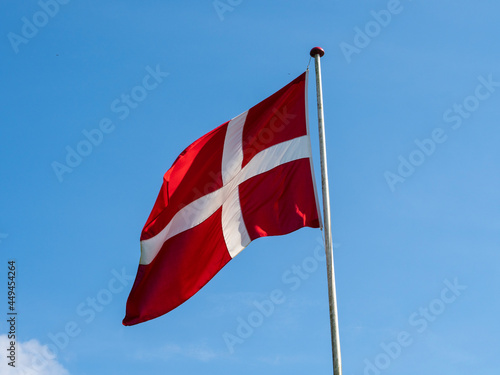 The Danish flag Dannebrog on a flagpole up against a blue sky with white clouds. Red flag with white cross. Denmark. 