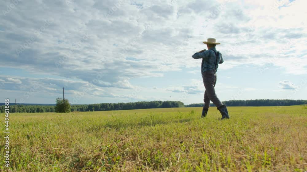 Farmer wearing straw hat, suspenders and rubber boots walking in mown field. Back view of gardener working in farmland. Man walks around plantation after farming work. Sunny day in countryside Farmer wearing straw hat, suspenders and rubber boots walking in mown field. Back view of gardener working in farmland. Man walks around plantation after farming work. Sunny day in countryside
