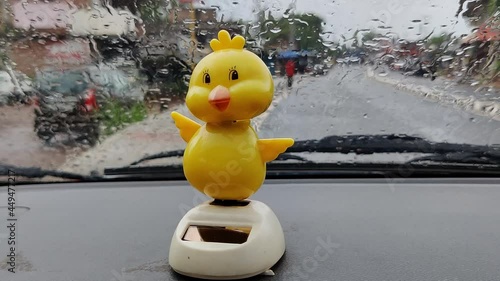 Yellow toy duck with its shaking head on top of the dashboard of a car parked on the side of the road in the rain