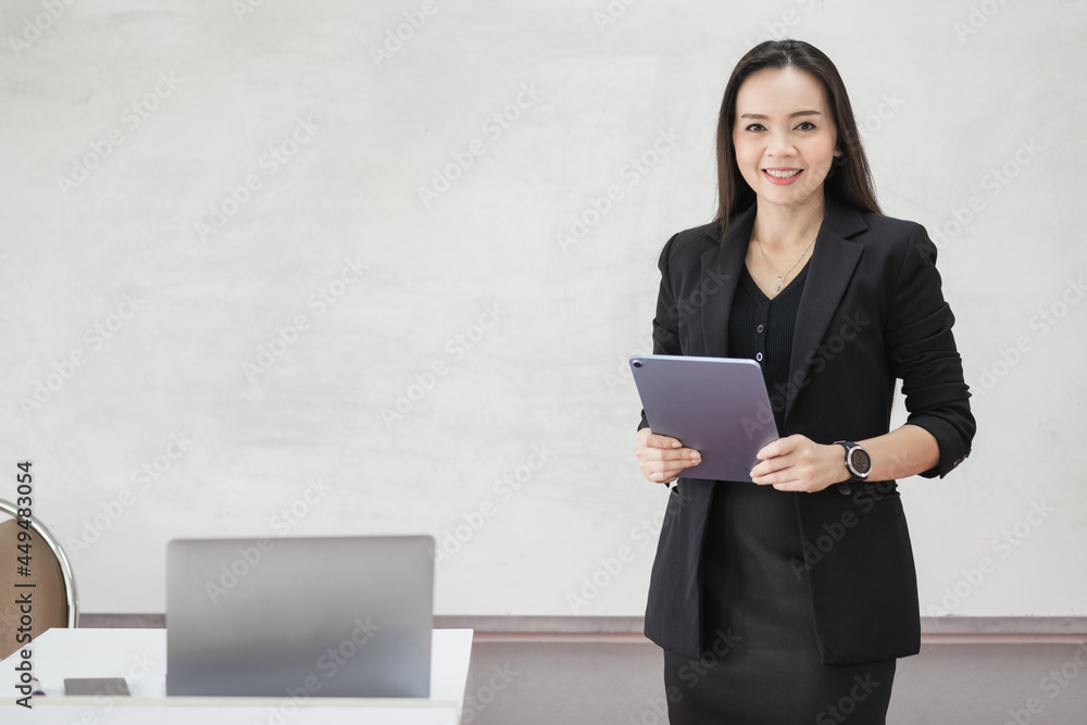 Stock photo portrait of a confident cheerful Asian woman teacher in a black business suit uniform with a digital tablet and laptop to teach modern language in the classroom Stock photo portrait of a confident cheerful Asian woman teacher in a black business suit uniform with a digital tablet and laptop to teach modern language in the classroom