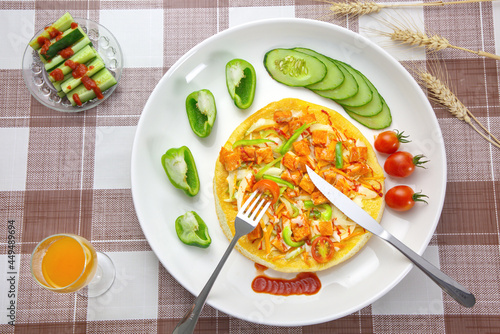 A self-made pizza in a white plate with cucumbers, green peppers, tomatoes, dipping sauce, juice; a kinfe, and a fork on a brown checkered table sheet; top view photo