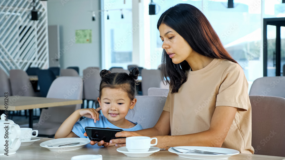 Lady shows daughter smartphone. Asian woman brunette and small kid girl watch video on black gadget sitting at restaurant table waiting for order close view Lady shows daughter smartphone. Asian woman brunette and small kid girl watch video on black gadget sitting at restaurant table waiting for order close view